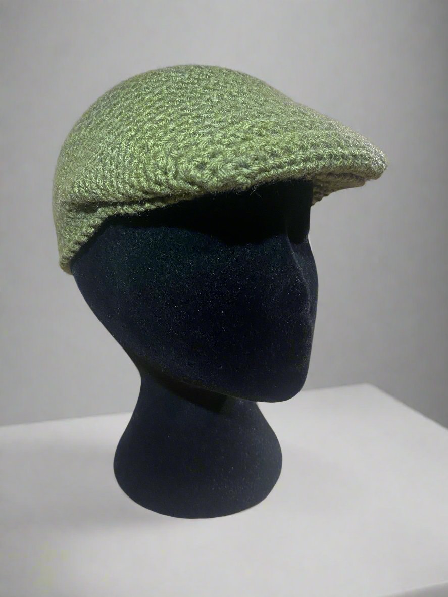 An olive crochet newsboy cap displayed on a mannequin head against a marble background.