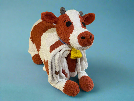 A crochet blanket with a cow design, featuring white body with brown spots, and a blue rim around the edge, displayed on a circular frame.
