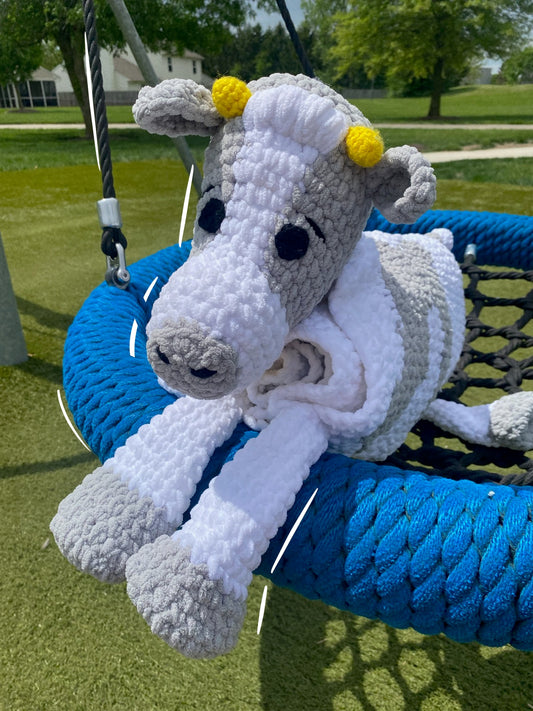 A crochet blanket with a cow design, featuring white body with gray and black spots, and a blue rim around the edge, displayed on a circular frame.