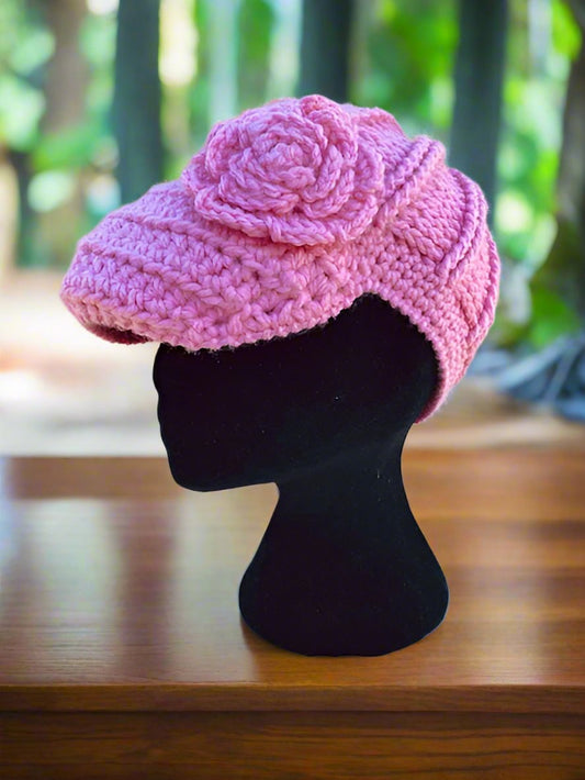 A handcrafted crochet hat in pink with a flower design, displayed on a mannequin head, set against a natural background.