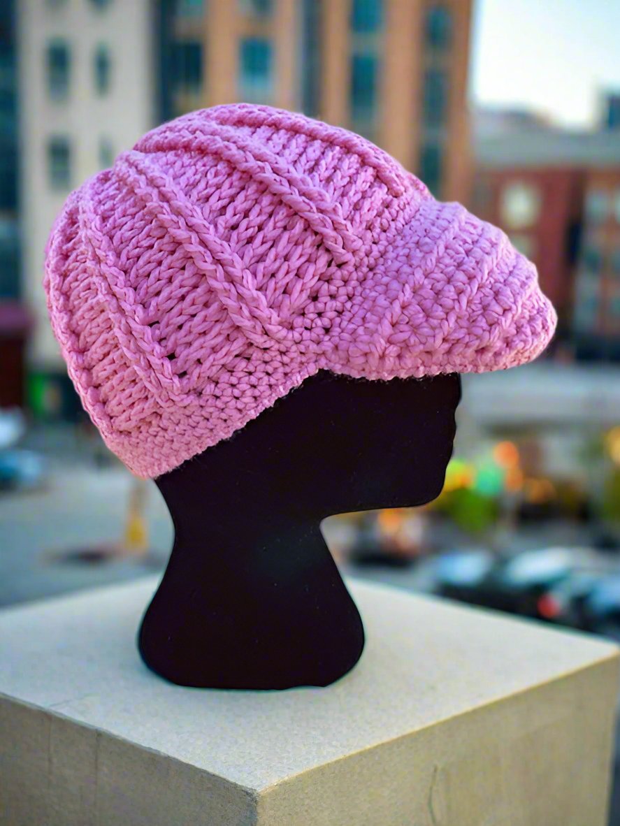 A handcrafted crochet hat in pink, displayed on a mannequin head, set against a city background.