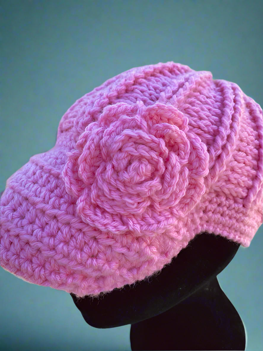 A handcrafted crochet hat in pink with a flower design, displayed on a mannequin head, set against a blue background