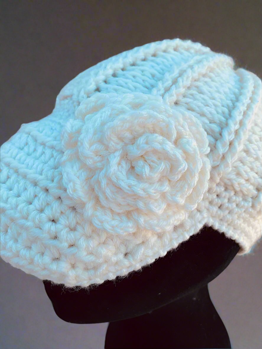 A handcrafted crochet hat in white with a flower design, displayed on a mannequin head, set against a gray background.