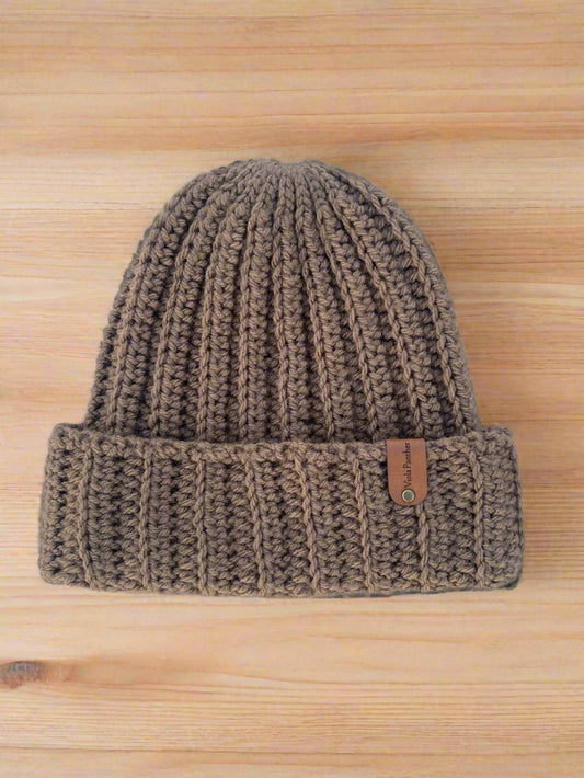 A brown, handmade crochet cuffed ribbed beanie with a brand tag on the brim, displayed on a wooden surface.