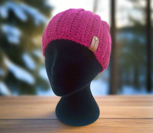 A pink handmade crochet rimless beanie displayed on a black mannequin head, set against a background with snow and trees.
