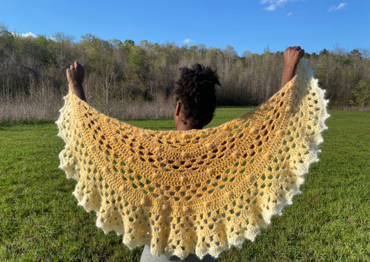 A person standing in an open field displaying a yellow and beige colored shawl with a patterned design spread out in front of them.