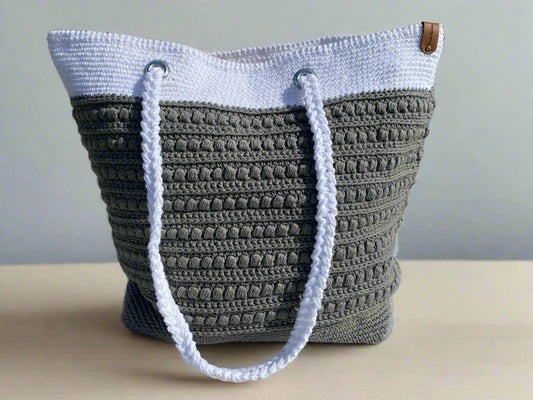 A crochet tote bag with two-tone design, featuring grey and white colors with braided handles and a faux leather tag.