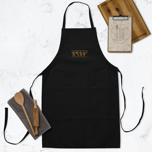 A black embroidered apron with front pockets and adjustable neck loop, displayed with a wooden spoon and a notebook.