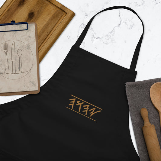 Close of of a black embroidered apron with front pockets and adjustable neck loop, displayed with a wooden spoon and a notebook.