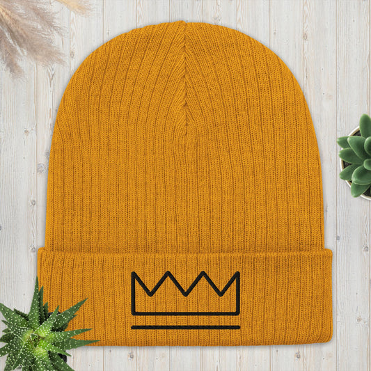 A yellow ribbed knit beanie with a black crown logo on the front, displayed on a wooden surface with a plant in the background.