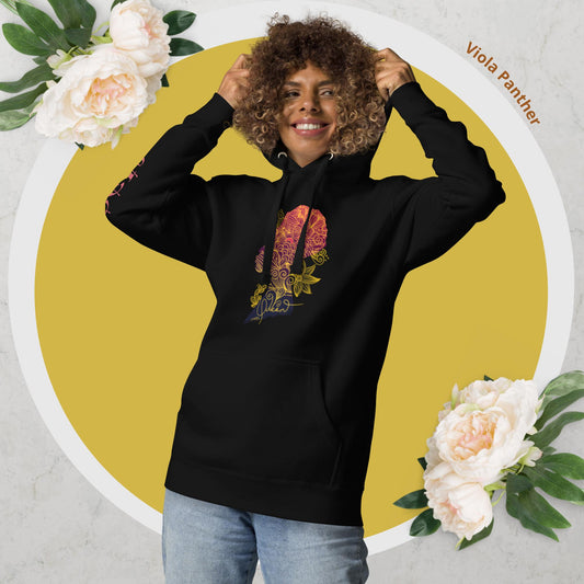 A woman smiling and posing with a black hoodie featuring a colorful floral design on the front. The hoodie is pulled up, revealing the design, and the woman has a flower in her hand.