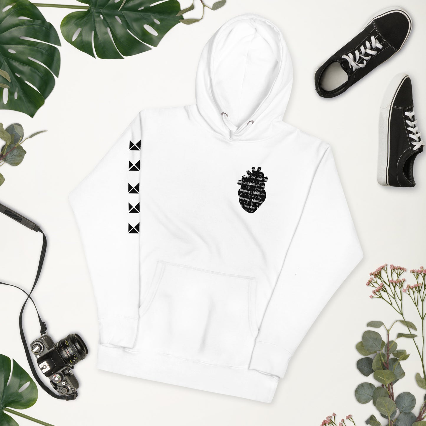A white unisex hoodie with a text design on the front, paired with black sneakers and a flat-brimmed hat, displayed against a background with green leaves and a camera.