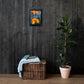 A canvas painting of an orange flower displayed on a wooden wall, with a potted plant and a wicker basket with a blue blanket in the foreground.